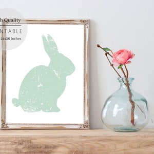 Puede incluir: Una impresi&oacute;n de arte imprimible con una silueta de conejo verde claro desgastada. La impresi&oacute;n est&aacute; enmarcada en un marco de madera blanco y se muestra en un estante de madera con un jarr&oacute;n de flores rosas.