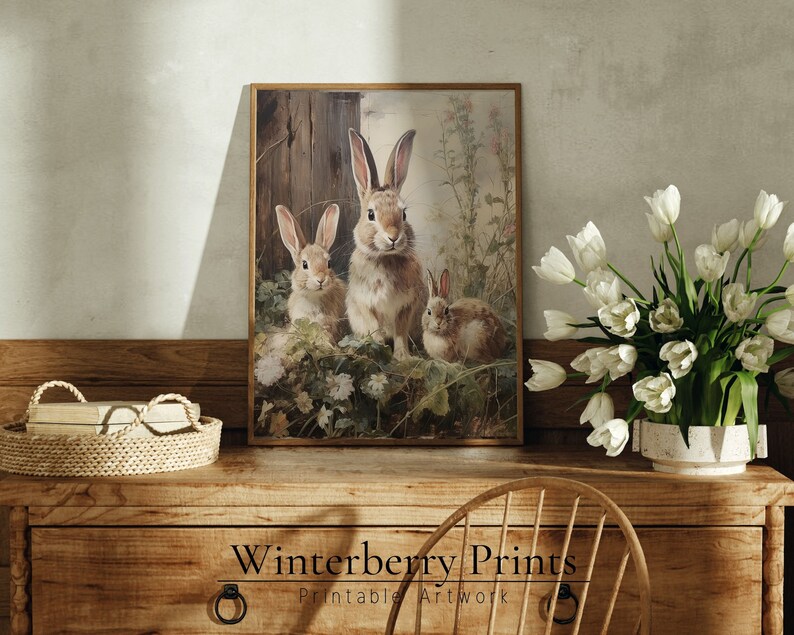May include: A vintage-style illustration of three brown rabbits in a field of flowers. The rabbits are sitting in a grassy area with a wooden fence in the background. The image is framed in a brown wooden frame.