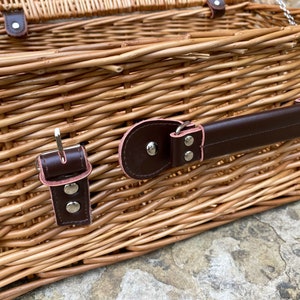 May include: A wicker picnic basket with a brown leather handle and two brown leather clasps with silver buckles. The basket is made of woven wicker and has a natural brown color.