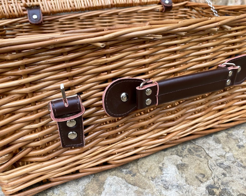 May include: A wicker picnic basket with a brown leather handle and two brown leather clasps with silver buckles. The basket is made of woven wicker and has a natural brown color.