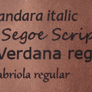 May include: Close-up of a brown leather surface with black text listing different font names and styles. The text reads: 1 - Candara italic, 2 - Segoe Script, 3 - Verdana regular, 4 - Gabriola regular.