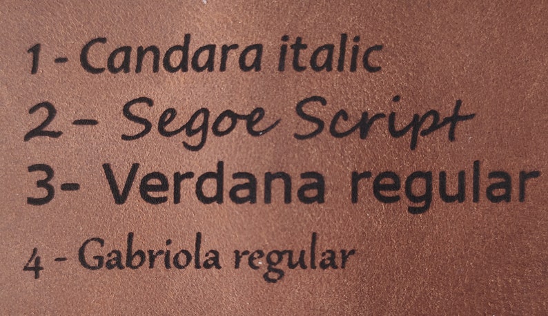 May include: Close-up of a brown leather surface with black text listing different font names and styles. The text reads: 1 - Candara italic, 2 - Segoe Script, 3 - Verdana regular, 4 - Gabriola regular.