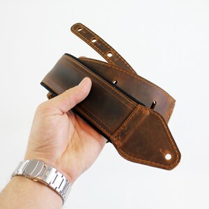 May include: A brown leather guitar strap with a black lining and stitching. The strap has a pointed end with a hole for attaching to a guitar.