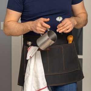 May include: A black canvas apron with multiple pockets, a white kitchen towel, a stainless steel milk frothing pitcher, a wooden spoon, and a thermometer.