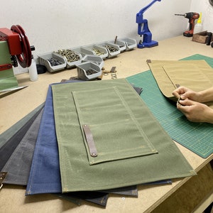 May include: Five different colored canvas work mats with leather handles and pockets. The mats are stacked on top of each other. A person is working on a tan mat, attaching a leather strap with a rivet.