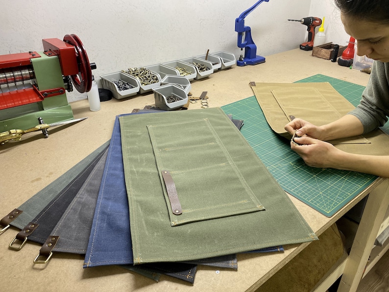 May include: Five different colored canvas work mats with leather handles and pockets. The mats are stacked on top of each other. A person is working on a tan mat, attaching a leather strap with a rivet.