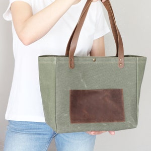 May include: A green canvas tote bag with brown leather handles and a brown leather patch pocket. The bag is being held by a person wearing a white t-shirt and blue jeans.