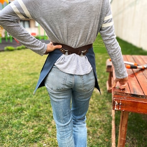 May include: A person wearing a grey long-sleeved shirt with white stripes on the sleeves, blue jeans, and a blue denim apron with a brown leather strap. The apron has a buckle closure and is worn over the shirt.