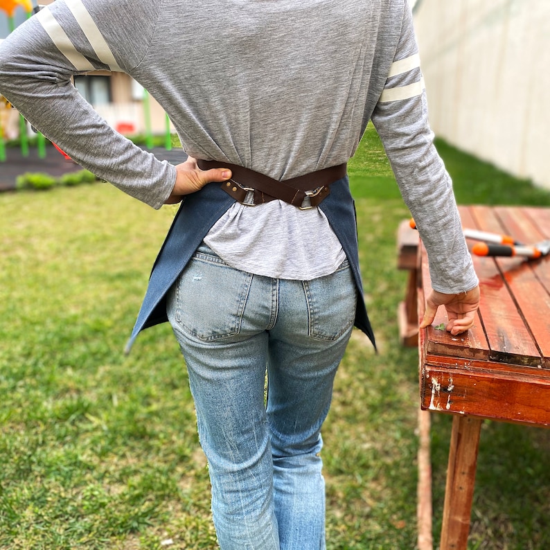May include: A person wearing a grey long-sleeved shirt with white stripes on the sleeves, blue jeans, and a blue denim apron with a brown leather strap. The apron has a buckle closure and is worn over the shirt.