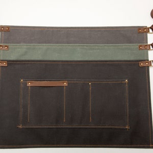 May include: Three brown canvas aprons with adjustable straps and a large pocket. The aprons are layered on top of each other, with the top apron being a dark gray, the middle apron being a light green, and the bottom apron being a dark brown.