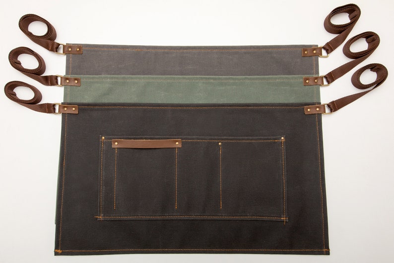 May include: Three brown canvas aprons with adjustable straps and a large pocket. The aprons are layered on top of each other, with the top apron being a dark gray, the middle apron being a light green, and the bottom apron being a dark brown.