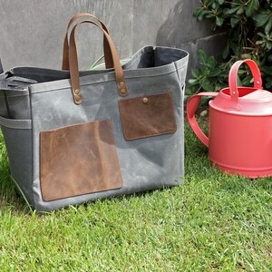 K&ouml;nnte beinhalten: Eine graue Canvas-Tragetasche mit braunen Lederelementen und Taschen. Die Tasche liegt auf gr&uuml;nem Gras neben einer roten Gie&szlig;kanne.