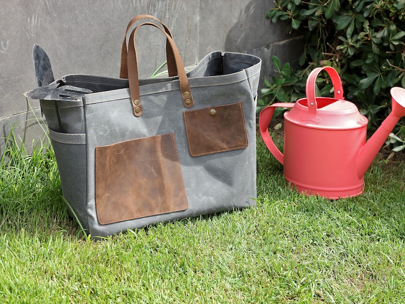 K&ouml;nnte beinhalten: Eine graue Canvas-Tragetasche mit braunen Lederelementen und Taschen. Die Tasche liegt auf gr&uuml;nem Gras neben einer roten Gie&szlig;kanne.