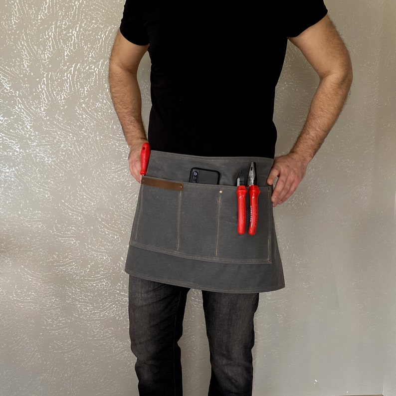 May include: A gray canvas tool apron with multiple pockets, including a large pocket for a phone or tablet. The apron has a brown leather strap and is worn by a person with red tools in the pockets.