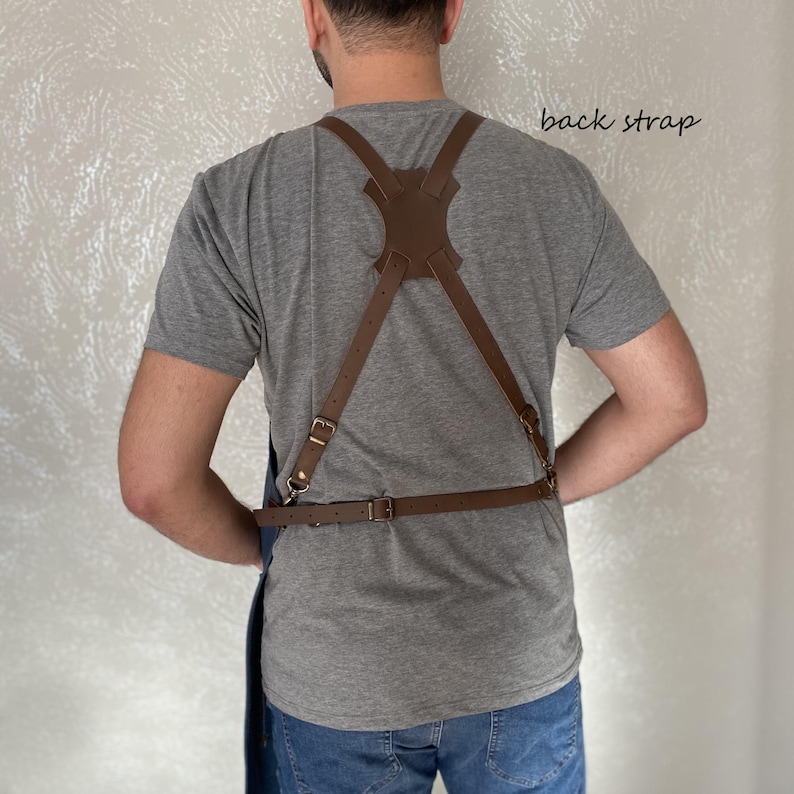 May include: A blue denim apron with brown leather straps and a back strap. The straps are adjustable and have brass buckles. The apron is worn by a person with a grey shirt and blue jeans.