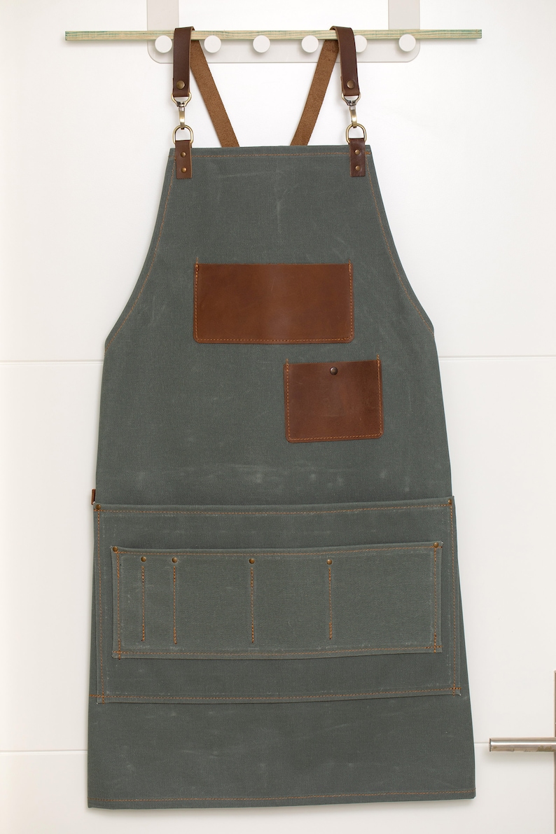 Op de afbeelding: Een grijze canvas schort met bruine leren banden en zakken. Het schort heeft een grote zak aan de voorkant met twee kleinere zakken erboven. Het schort hangt aan een haak.
