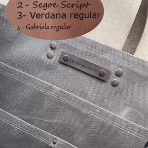 K&ouml;nnte beinhalten: Eine graue Canvas-Tragetasche mit einem braunen Ledergriff und einem braunen Leder-Patch mit der Aufschrift "Personalisierung". Die Tasche hat mehrere Taschen und ist mit Metallnieten gesichert.