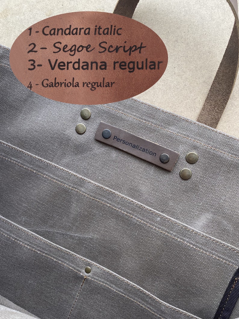 K&ouml;nnte beinhalten: Eine graue Canvas-Tragetasche mit einem braunen Ledergriff und einem braunen Leder-Patch mit der Aufschrift "Personalisierung". Die Tasche hat mehrere Taschen und ist mit Metallnieten gesichert.