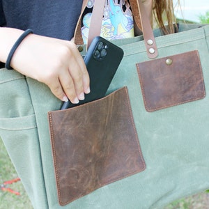 K&ouml;nnte beinhalten: Eine gr&uuml;ne Canvas-Tragetasche mit zwei braunen Ledertaschen und einem braunen Lederriemen. Ein schwarzes Telefon wird in die gr&ouml;&szlig;ere Tasche gelegt.