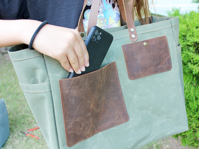 K&ouml;nnte beinhalten: Eine gr&uuml;ne Canvas-Tragetasche mit zwei braunen Ledertaschen und einem braunen Lederriemen. Ein schwarzes Telefon wird in die gr&ouml;&szlig;ere Tasche gelegt.