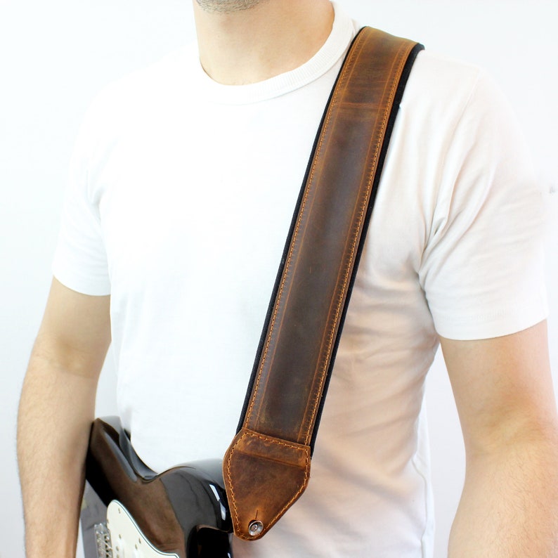 May include: A brown leather guitar strap with a dark brown stitching detail. The strap is worn over the shoulder of a person wearing a white t-shirt.