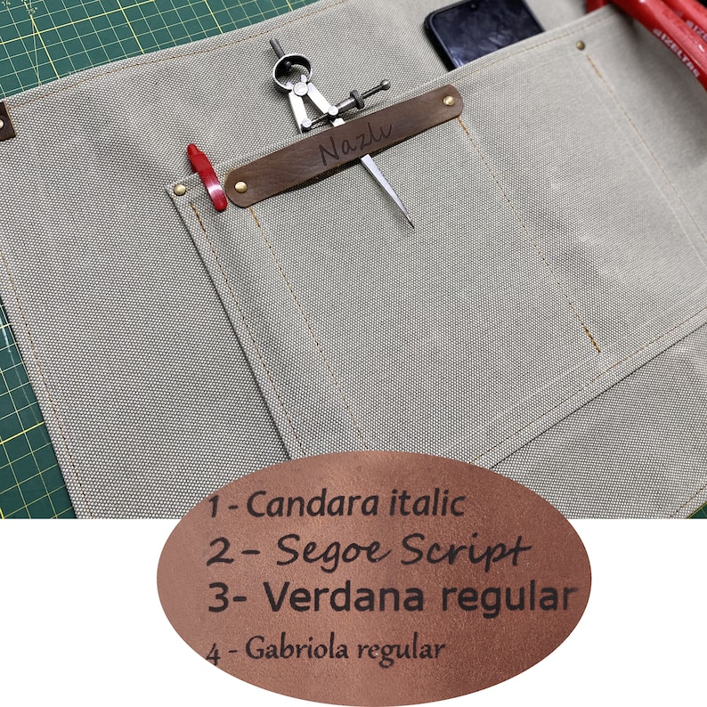 May include: A beige canvas apron with multiple pockets, including one with a brown leather patch that says "Nazlu" in cursive. The apron has a compass and a red pencil sticking out of the pockets. A brown leather patch on the bottom of the apron lists four different fonts: Candara italic, Segoe Script, Verdana regular, and Gabriola regular.