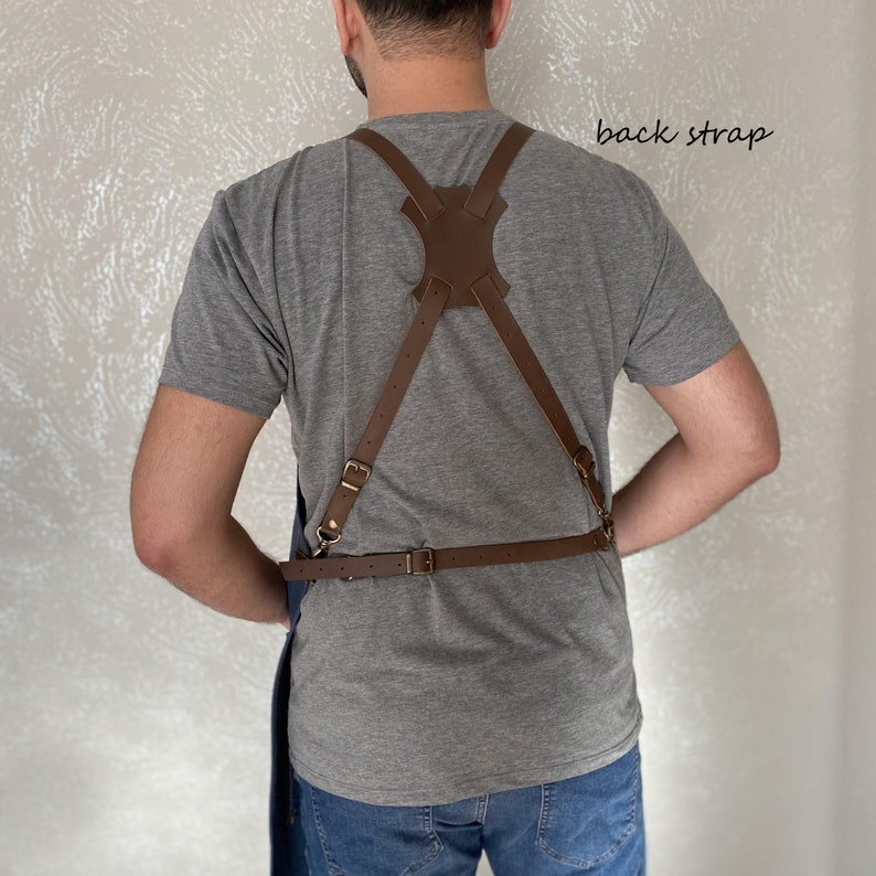 Op de afbeelding: Een blauwe denim schort met bruine leren banden en een rugband. De banden zijn verstelbaar en hebben messing gespen. Het schort wordt gedragen door een persoon met een grijs shirt en een blauwe jeans.