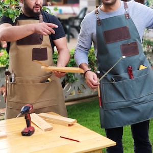 May include: Two people wearing brown and green canvas aprons with leather pockets and tool loops. The aprons have multiple pockets and loops for carrying tools. The person on the left is holding a piece of wood. The person on the right is holding a metal tool.