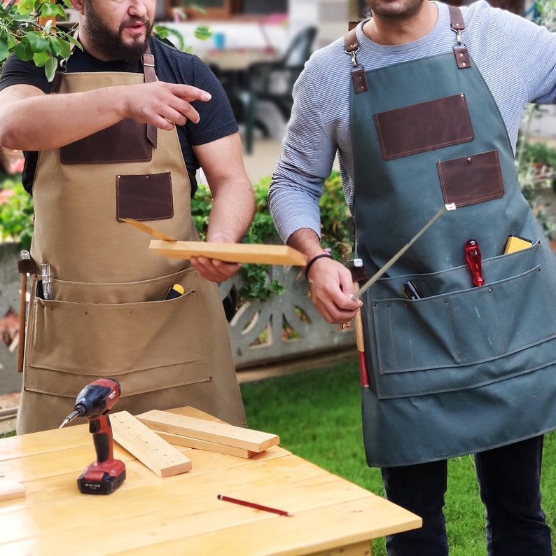 May include: Two people wearing brown and green canvas aprons with leather pockets and tool loops. The aprons have multiple pockets and loops for carrying tools. The person on the left is holding a piece of wood. The person on the right is holding a metal tool.