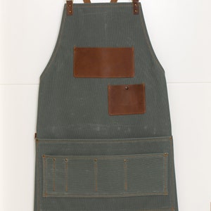 May include: A gray canvas apron with brown leather straps and pockets. The apron has a large pocket on the front with two smaller pockets above it. The apron is hanging on a hook.