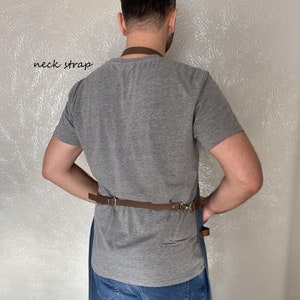 Op de afbeelding: Een persoon draagt een grijs T-shirt en een blauwe jeans met een bruine leren schort. Het schort heeft een bruine leren nekband en een bruine leren tailleband met een metalen gesp. De tekst "neck strap" is in zwart op een witte achtergrond geschreven.