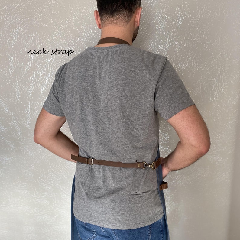 Op de afbeelding: Een persoon draagt een grijs T-shirt en een blauwe jeans met een bruine leren schort. Het schort heeft een bruine leren nekband en een bruine leren tailleband met een metalen gesp. De tekst "neck strap" is in zwart op een witte achtergrond geschreven.
