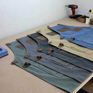 Op de afbeelding: Vijf schorten van canvas in verschillende kleuren, waaronder groen, grijs, blauw, beige en bruin, met leren banden en zakken. De schorten liggen op een houten tafel.