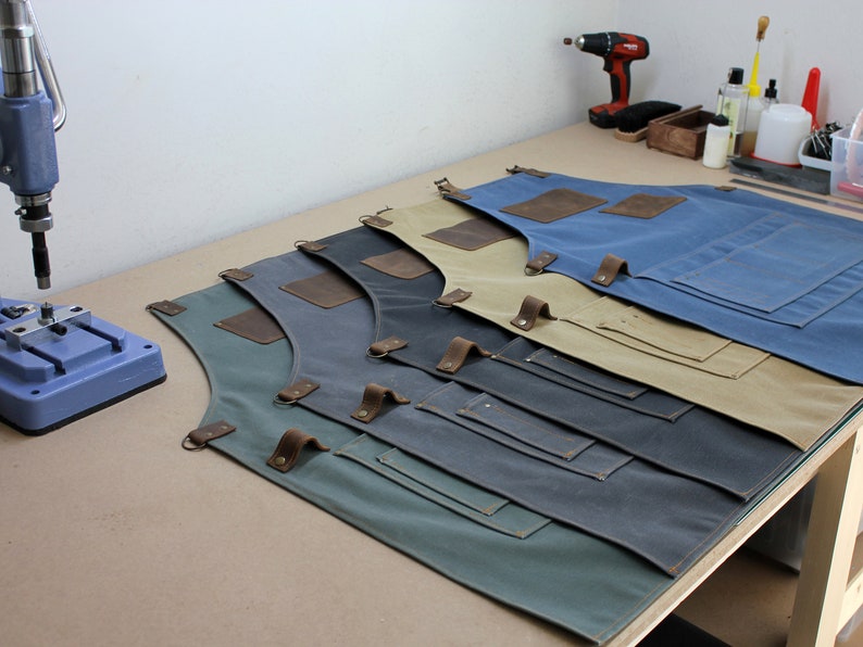 Op de afbeelding: Vijf schorten van canvas in verschillende kleuren, waaronder groen, grijs, blauw, beige en bruin, met leren banden en zakken. De schorten liggen op een houten tafel.