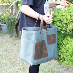 K&ouml;nnte beinhalten: Eine gro&szlig;e, gr&uuml;ne Canvas-Tragetasche mit braunen Lederelementen und Taschen. Die Tasche hat zwei lange Henkel und ist aus strapazierf&auml;higem Material.