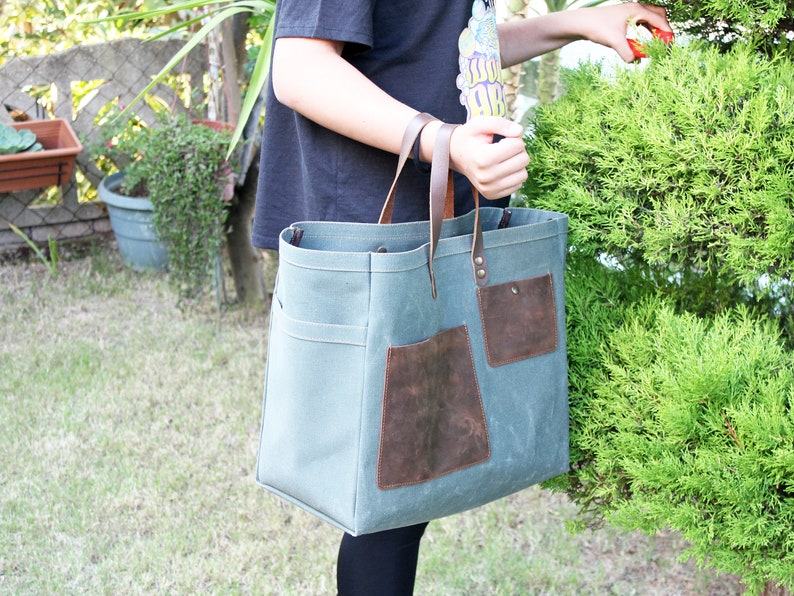 K&ouml;nnte beinhalten: Eine gro&szlig;e, gr&uuml;ne Canvas-Tragetasche mit braunen Lederelementen und Taschen. Die Tasche hat zwei lange Henkel und ist aus strapazierf&auml;higem Material.