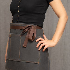 May include: A gray canvas apron with brown straps and a brown ribbon tied at the waist. The apron has two large pockets on the front and is designed for use in a kitchen or workshop.