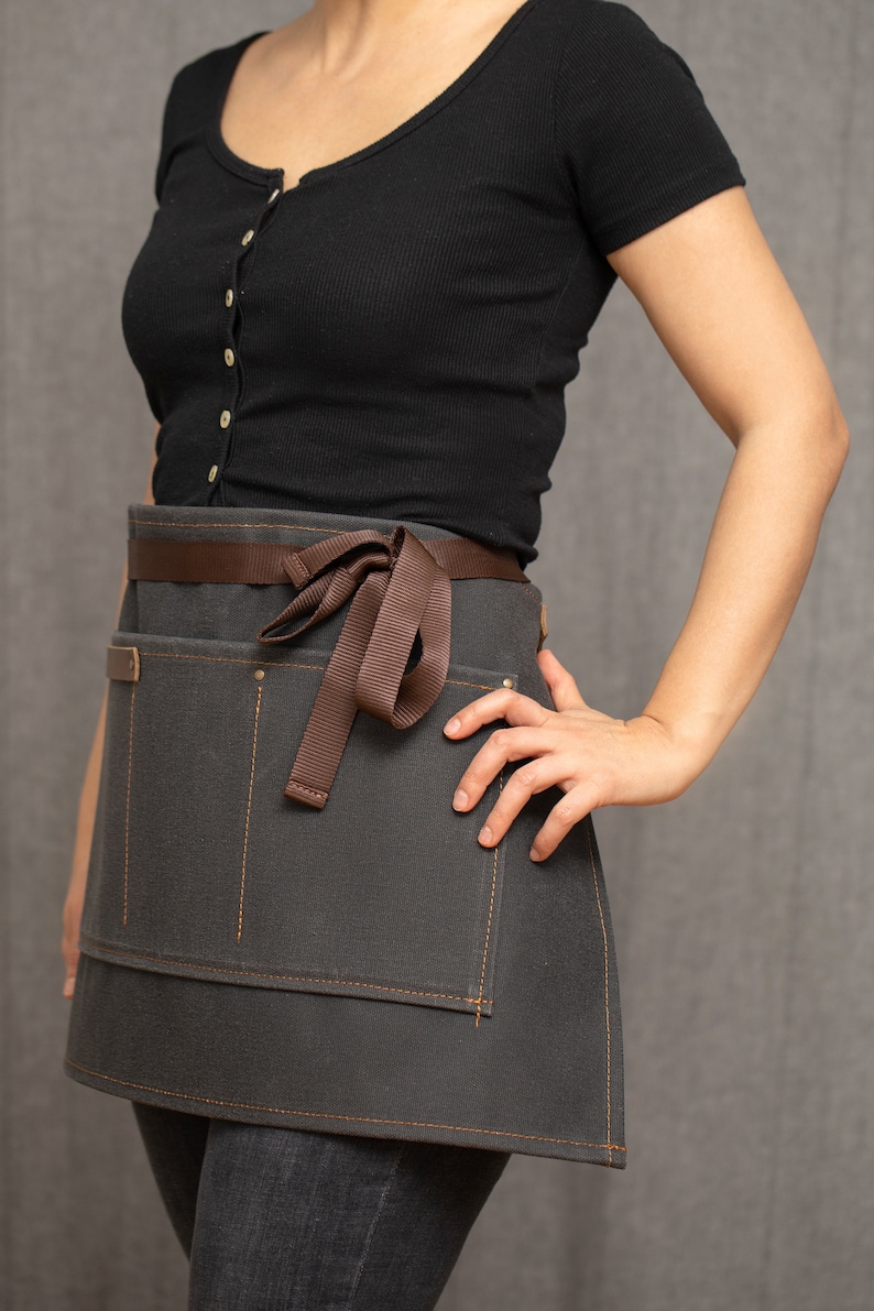 May include: A gray canvas apron with brown straps and a brown ribbon tied at the waist. The apron has two large pockets on the front and is designed for use in a kitchen or workshop.
