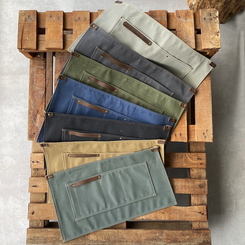 May include: A stack of eight canvas aprons in different colors, including green, blue, brown, and gray. Each apron has a leather strap and multiple pockets.