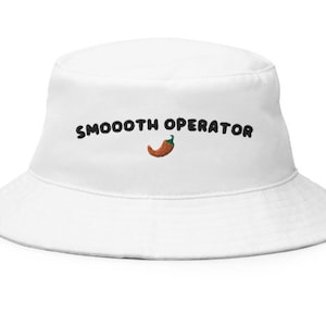 Może przedstawiać: Biały kapelusz typu bucket z haftowanym napisem "SMOOOOTH OPERATOR" i ilustracją papryczki chili. Kapelusz ma szerokie rondo i swobodny styl, idealny na słoneczne dni i do aktywności na świeżym powietrzu.