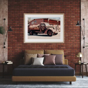 Puede incluir: Un cami&oacute;n rojo oxidado vintage con un tanque de agua es el punto focal de una fotograf&iacute;a enmarcada que cuelga de una pared de ladrillo en un dormitorio. El cami&oacute;n est&aacute; estacionado frente a un edificio con una puerta de madera. El dormitorio cuenta con una cama con un colch&oacute;n azul oscuro y una cabecera marr&oacute;n, junto con algunas almohadas. Hay dos mesitas de noche con l&aacute;mparas y plantas.