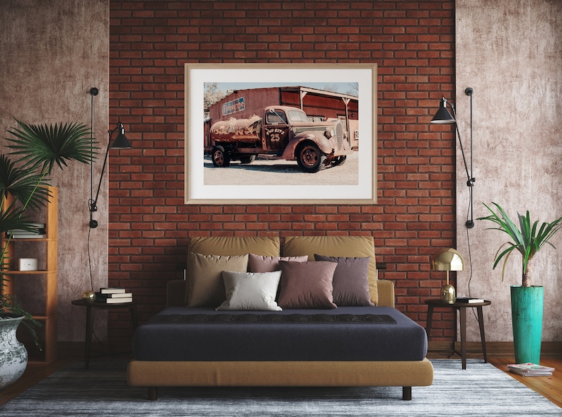 Puede incluir: Un cami&oacute;n rojo oxidado vintage con un tanque de agua es el punto focal de una fotograf&iacute;a enmarcada que cuelga de una pared de ladrillo en un dormitorio. El cami&oacute;n est&aacute; estacionado frente a un edificio con una puerta de madera. El dormitorio cuenta con una cama con un colch&oacute;n azul oscuro y una cabecera marr&oacute;n, junto con algunas almohadas. Hay dos mesitas de noche con l&aacute;mparas y plantas.