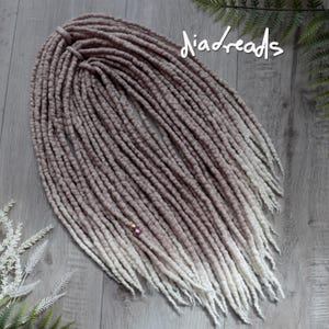 May include: A set of synthetic dreadlocks featuring a soft taupe to white ombre color transition. The individual strands have a textured, matte appearance and are adorned with two small, metallic beads. The text "diadreads" is visible in white.