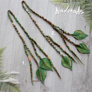 May include: Two long hair extensions labeled 1 and 2, featuring braided green and brown fibers adorned with small beads and felted leaf shapes at the ends. The extensions incorporate white thread accents and are arranged on a light wood surface.