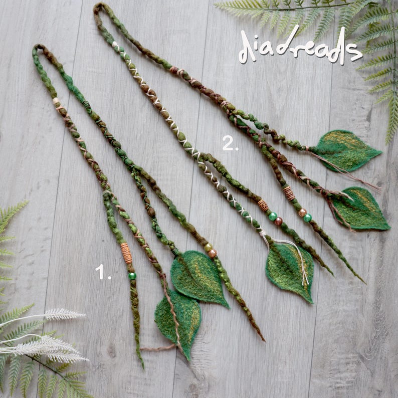 May include: Two long hair extensions labeled 1 and 2, featuring braided green and brown fibers adorned with small beads and felted leaf shapes at the ends. The extensions incorporate white thread accents and are arranged on a light wood surface.