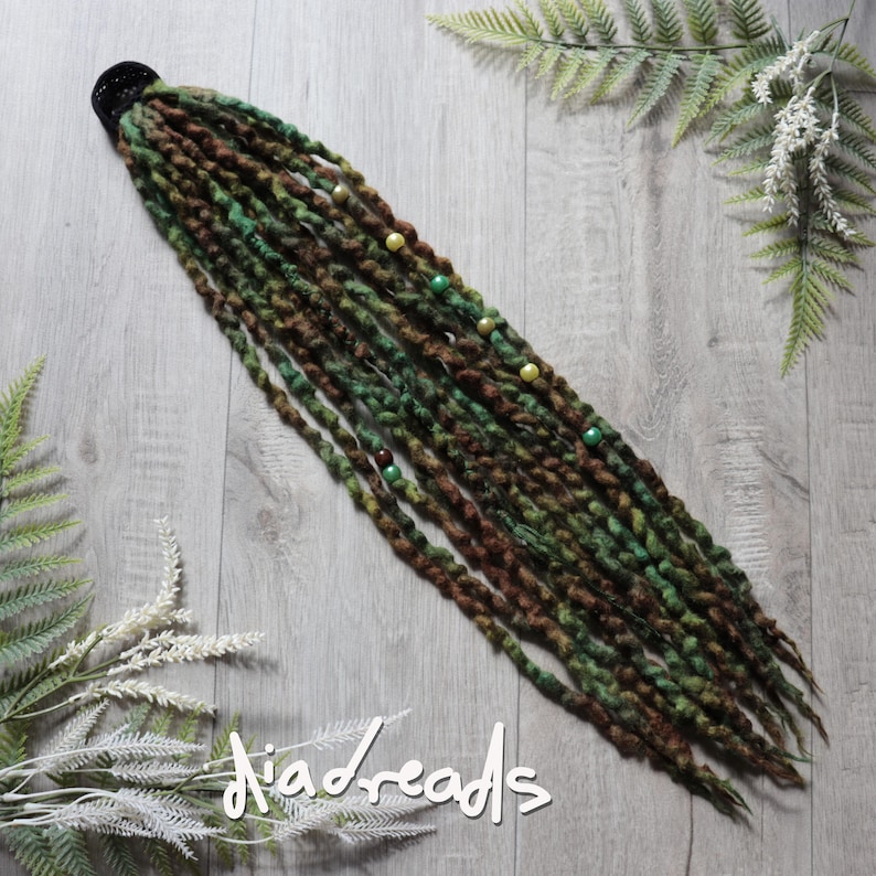 May include: A set of synthetic dreadlocks in shades of forest green and dark brown, attached to a black hair tie. The dreadlocks feature a twisted texture and are accented with small, round beads in yellow, green, and brown colors.