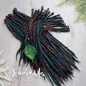 May include: A bundle of handmade dreadlocks in green, blue, and purple hues. A green leaf-shaped decoration is attached to the dreadlocks. The text "diadreads" is written in a cursive font across the bottom of the image.