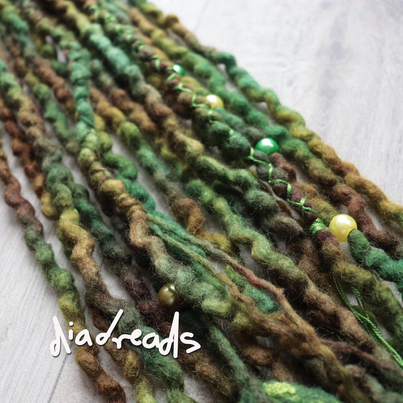 May include: A collection of textured synthetic dreadlocks in shades of forest green, olive, and dark brown. The strands feature a wavy, felted appearance with decorative round beads in cream and green, and the text diadreads is visible.
