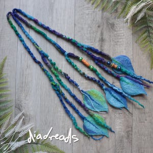 May include: A set of four long, braided hair extensions in shades of blue, green, and purple. Each braid terminates in a felted leaf shape. The braids are accented with small green, blue, and orange beads. The text "diadreads" is visible at the bottom.