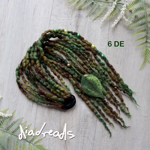 May include: A set of synthetic dreadlocks in shades of forest green, olive, and brown, featuring a black circular attachment point. The dreads are accented with small green and yellow beads and a single felted green leaf. Text reads 6 DE.